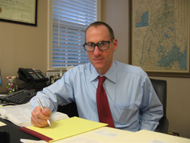 Adam Bernstein wearing black rimmed glasses, a blue long-sleeved shirt, and red tie holding a pen in hand with a yellow notepad in front of him, looking at the camera.
